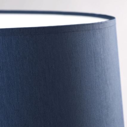 Tmavomodrý tienidlo s bielou vnútornou časťou stojí osamote na svetlom pozadí. Tienidlo je valcového tvaru, vyrobené z látky s jemnou textúrou.