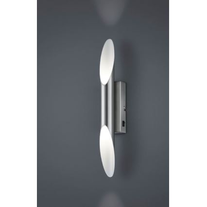 Dvojité nástenné svietidlo z nehrdzavejúcej ocele s oválnymi difúzormi svieti na tmavošedej stene. Svietidlo je pripevnené na stenu pomocou štvorcovej základne so spínačom.