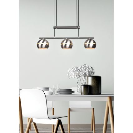 Trojramenná závesná lampa s kovovými guľovými tienidlami visí nad jedálenským stolom. Lampa je zavesená na tyči. Stôl je biely s drevenými nohami, okolo neho sú biele stoličky. Na stole je váza s kvetmi.