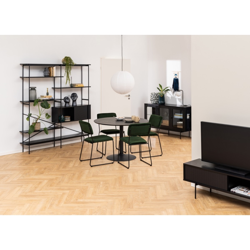 Jedálenský stôl s okrúhlou doskou a štyrmi tmavozelenými stoličkami stojí v miestnosti s hnedou parketou. V pozadí je vysoká čierna polica a televízor na čiernom stolíku. Miestnosť je zariadená v modernom štýle.