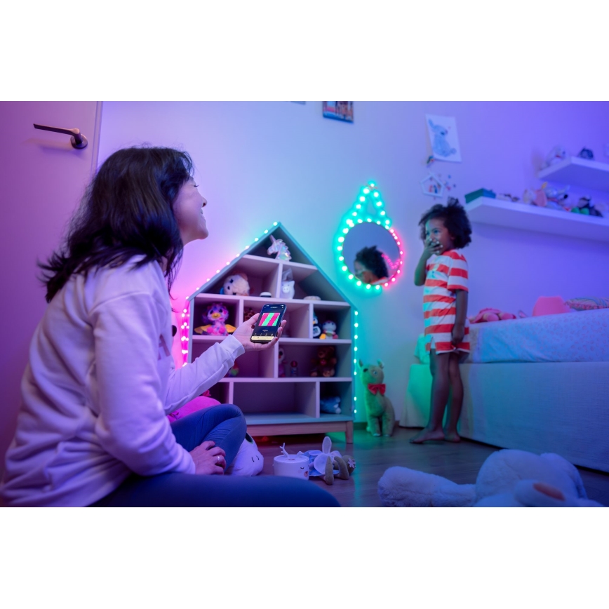 Žena v detskej izbe ovláda pomocou mobilného telefónu farebné LED osvetlenie. Dieťa stojí v pozadí. Osvetlenie je umiestnené na poličke v tvare domčeka a okolo zrkadla.