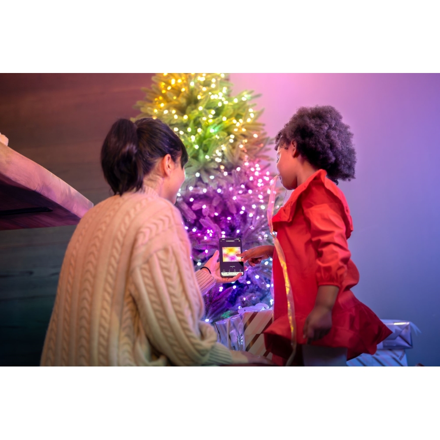 Dve ženy, dospelá a dievčatko, stoja pred vianočným stromčekom s farebnými svetlami. Dospelá žena drží telefón, na ktorom je zobrazený obrázok, a dievčatko ukazuje na displej. V pozadí sú vianočné darčeky.