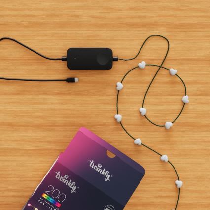 Svetelný reťaz s LED diódami v tvare srdiečok je pripojený k čiernemu adaptéru. Adaptér je položený na drevenom povrchu vedľa krabice s nápisom „twinkly™ 200 MULTICOLOR APP CONTROLLED RGB LED“.