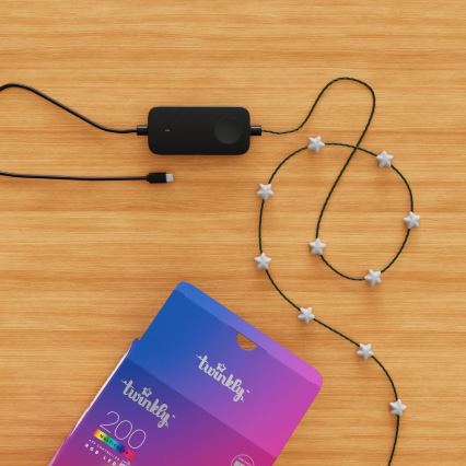 Svietiaca šnúra s hviezdami je pripojená k čiernemu ovládaču. Ovládač je napojený na USB kábel. Všetko leží na drevenom povrchu, vedľa otvorenej krabice s nápisom „twinkly™ 200 MULTICOLOR APP CONTROLLED RGB LED“.