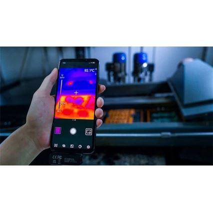 Mobilný telefón sníma termovíziu objektu. Na displeji sú zobrazené teploty v stupňoch Celzia (92,1 °C, 38,2 °C, 222,4 °C). Na pozadí je priemyselné zariadenie.