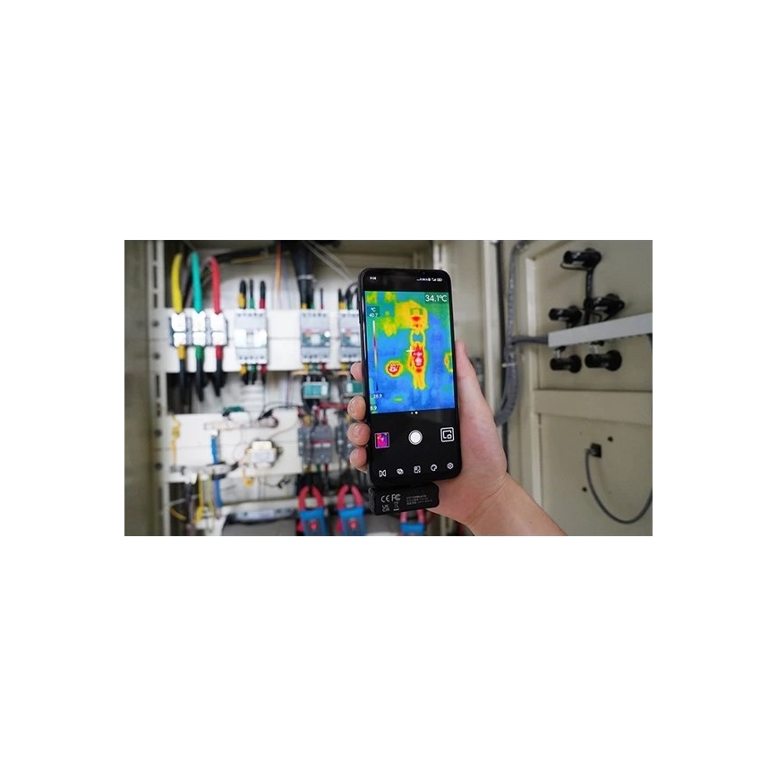 Mobilný telefón sníma termovíziu elektrického rozvádzača. Na displeji je zobrazená teplotná mapa s hodnotami od 28.5 °C do 40.7 °C a maximálnou teplotou 34.1 °C. Rozvádzač obsahuje rôzne elektrické komponenty a káble.