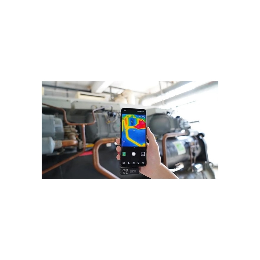 Mobilný telefón sníma termografický obraz technickej inštalácie. Na displeji je vidieť teplotné rozdiely v červeno-žltých a modrých odtieňoch, s vyznačenou teplotou 33.4°C. Na pozadí je priemyselná klimatizačná jednotka s potrubím.