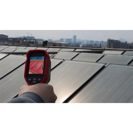 Termokamera sníma teplotu solárnych panelov na streche budovy. Na displeji termokamery je zobrazená teplotná mapa panelov s údajom Max 55,0 °C. V pozadí je vidieť mesto.