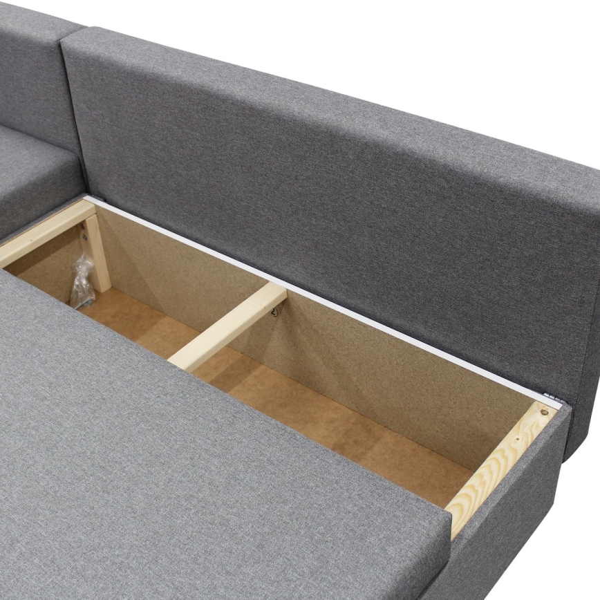 Rohový gauč s otvoreným úložným priestorom. Vnútorná konštrukcia z dreva je viditeľná. Sivá látková poťah.