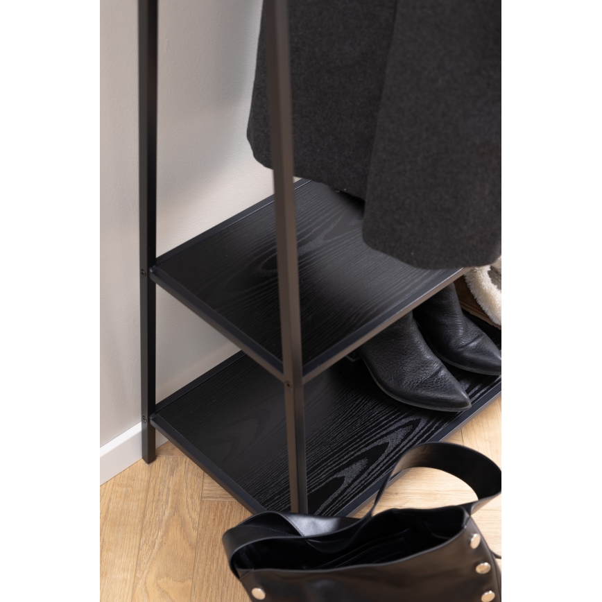 Čierny kovový stojan na oblečenie s tromi tmavými drevenými policami; na ňom visí kabát, sú na ňom topánky a taška. Stojan stojí na svetlej drevenej podlahe pri stene.