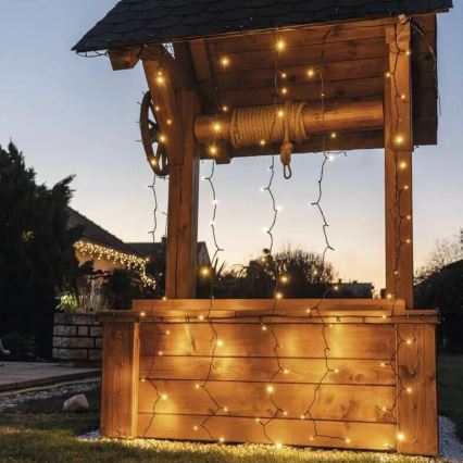 Drevený studničný domček je ozdobený svetelnými reťazami s teplými žiarovkami. Reťaze visia z vrchu a po stranách domu. Na pozadí sa nachádza záhrada a domy.