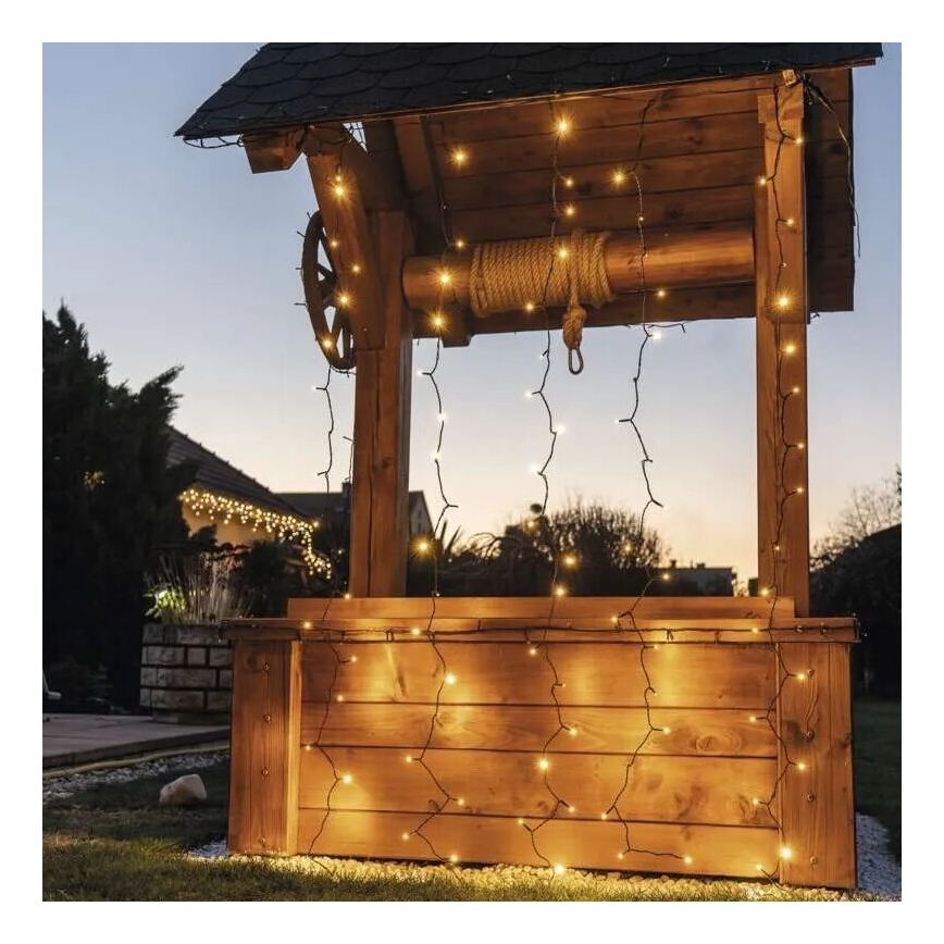 Drevený studničný domček je ozdobený svetelnými reťazami s teplými žiarovkami. Reťaze visia z vrchu a po stranách domu. Na pozadí sa nachádza záhrada a domy.