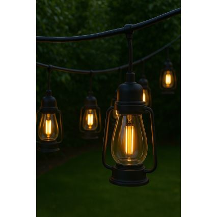 Svetlá v tvare lampášov svietia v exteriéri. Závesné lampáše s teplým žltým svetlom sú zavesené na čiernom kábli. Okolie tvorí tmavozelený trávnik a pozadie s rozmazanou zeleňou.