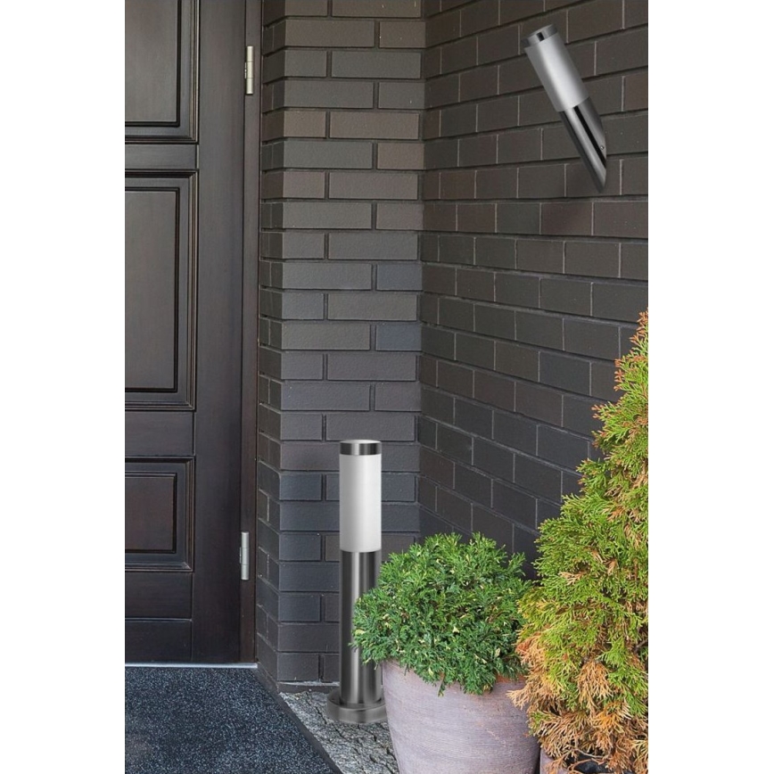 Stĺpová lampa stojí pri tmavohnedej dverách, vedľa tmavošedej tehlovej steny. Vedľa nej sú v kvetináčoch kríky. Na stene je umiestnená nástenná lampa.