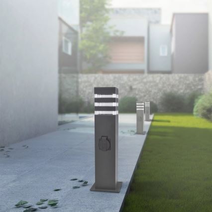 Svietiaca záhradná lampa s integrovanou zásuvkou stojí na chodníku. Lampa svieti. V pozadí je vidieť záhradu a modernú budovu.