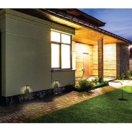 Dve vonkajšie nástenné svietidlá osvetľujú chodník pred domom. Svietidlá sú umiestnené na vonkajšej stene domu v noci. Dom má drevené obloženie a osvetlené okno.