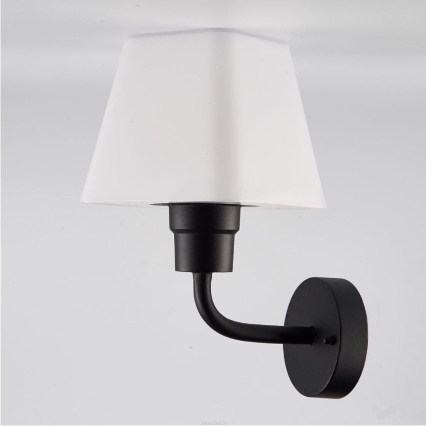 Nástenná lampa s bielym štvorcovým tienidlom je pripevnená k stene. Čierna kovová konštrukcia drží tienidlo a je pripevnená k okrúhlemu nástennému držiaku.