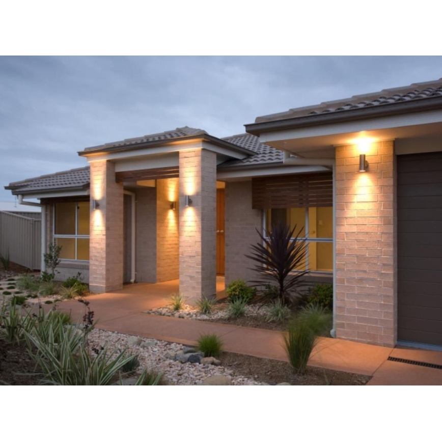 Vonkajšie nástenné svietidlá osvetľujú vstup do rodinného domu s hnedou strechou a svetlohnedými tehlovými stenami. Svietidlá sú umiestnené na stĺpikoch po oboch stranách vstupu. Pred domom sa nachádza chodník a záhradná úprava.