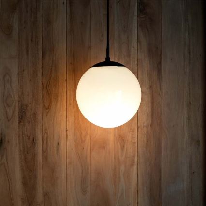 Guľová lampa svieti. Je zavesená na čiernom káble pred drevenou stenou.