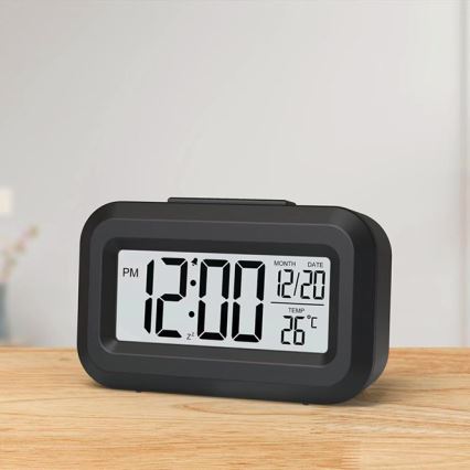 Digitálny budík zobrazuje čas 12:00 PM, dátum 12/20 a teplotu 26°C. Stojí na drevenom povrchu.
