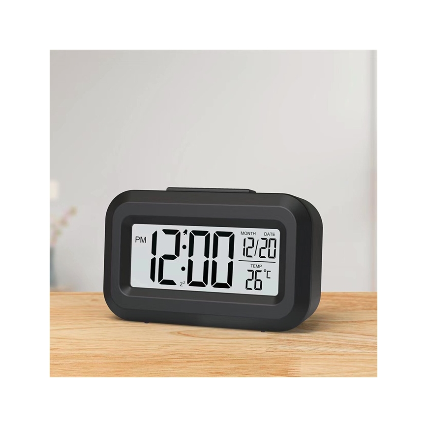 Digitálny budík zobrazuje čas 12:00 PM, dátum 12/20 a teplotu 26°C. Stojí na drevenom povrchu.