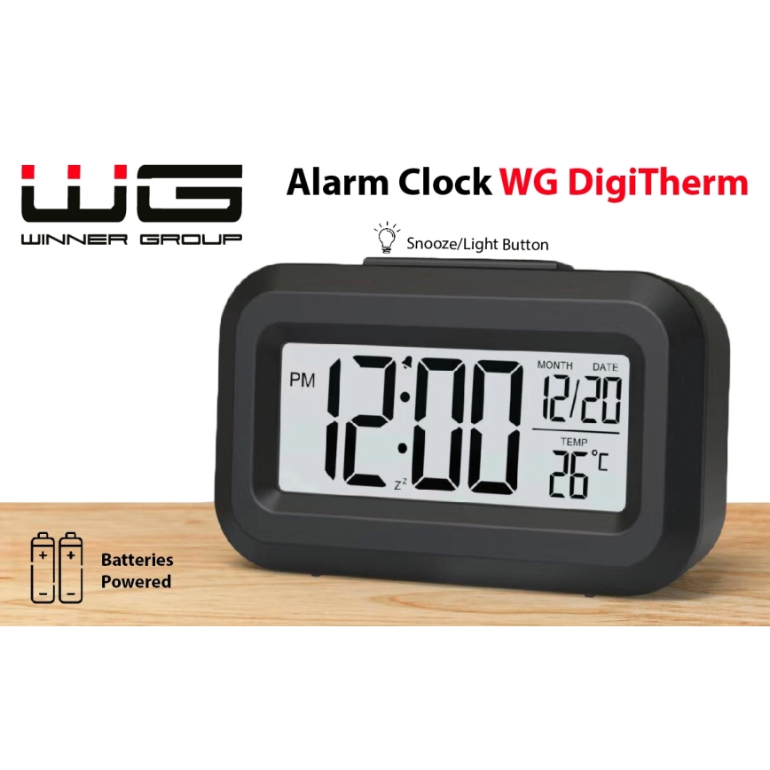 Čierny digitálny budík WG DigiTherm zobrazuje čas 12:00, dátum 12/20 a teplotu 26 °C. Je napájaný batériami a má tlačidlo pre odloženie budíka a podsvietenie. Stojí na drevenom povrchu.