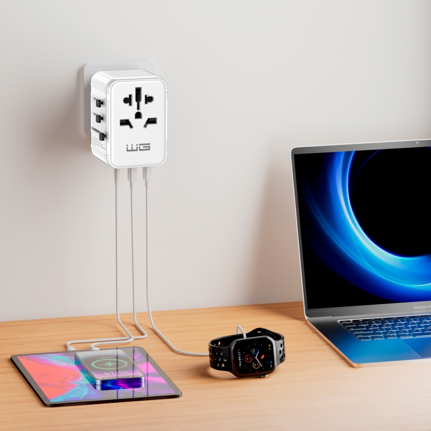 Biely cestovný adaptér nabíja tablet, hodinky Apple Watch a laptop. Adaptér je zapojený do steny. Všetky zariadenia sú položené na drevenom stole.