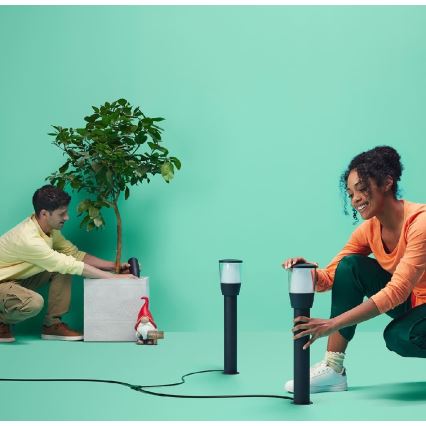 Dvaja ľudia inštalujú záhradné svietidlá na zelenej ploche. Muž sadí stromček do kvetináča. Žena montuje svietidlá pripojené káblom.