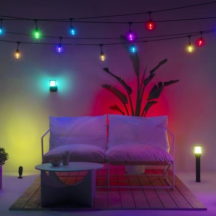 Dvojsedačka s vankúšmi je osvetlená farebnými svetlami. Svetlá sú umiestnené na stene, na stole a v okolí sedačky. Rastlina v črepníku sa nachádza za sedačkou. Všetko je umiestnené na drevenej terase.