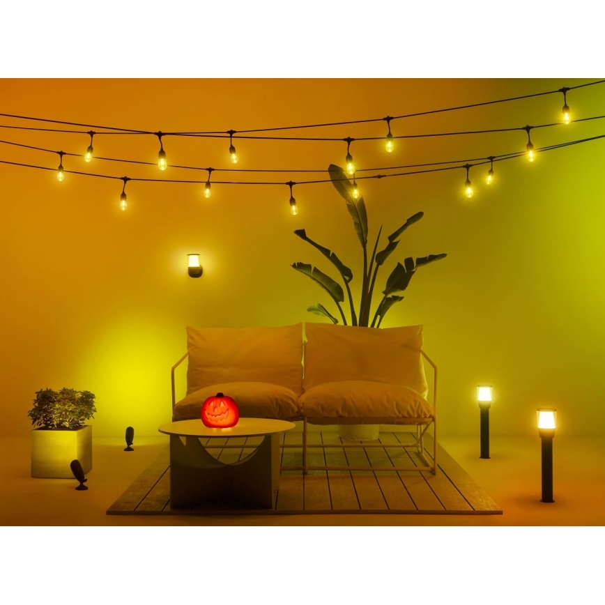 Svetlá svietia na terase s bielou sedačkou a okrasnou tekvicou na nízkom stolíku. Rastlina v kvetináči a niekoľko vonkajších svietidiel dopĺňajú scénu s prevahou teplej žltej farby.