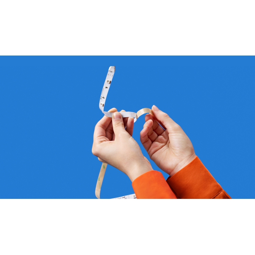 Dve ruky v oranžových rukávoch odstraňujú ochrannú fóliu z LED pásika. Pásik je biely s malými LED diódami. Pozadie je modré.