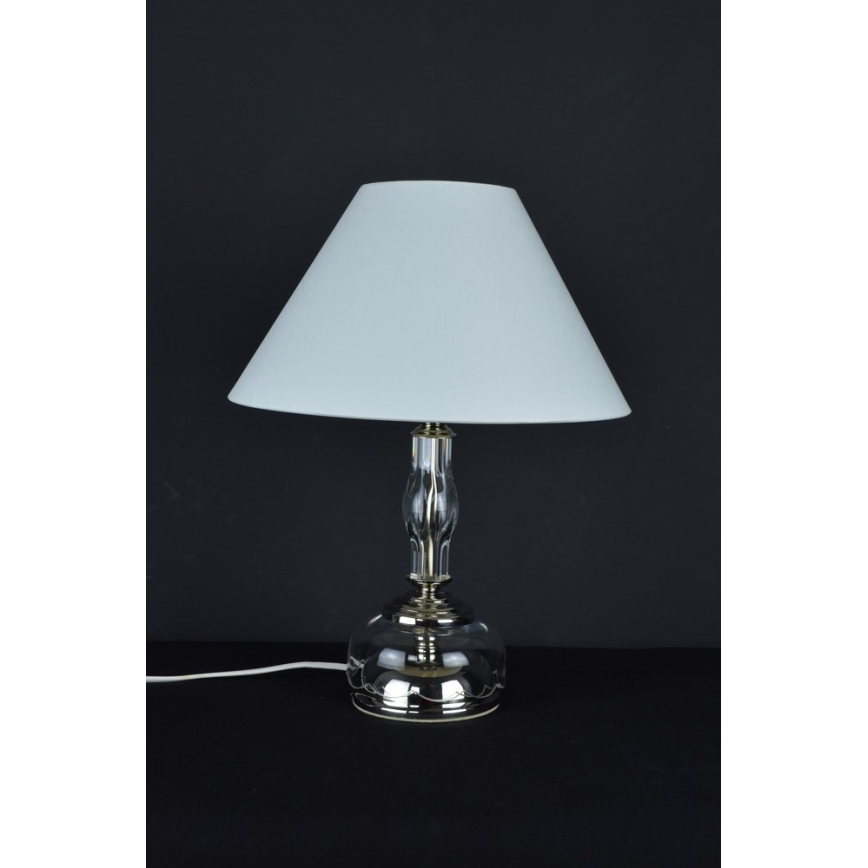 Stolová lampa s bielym tienidlom stojí na tmavom povrchu. Lampa má priehľadný stredový diel a kovový podstavec. Viditeľný je aj pripájací kábel.
