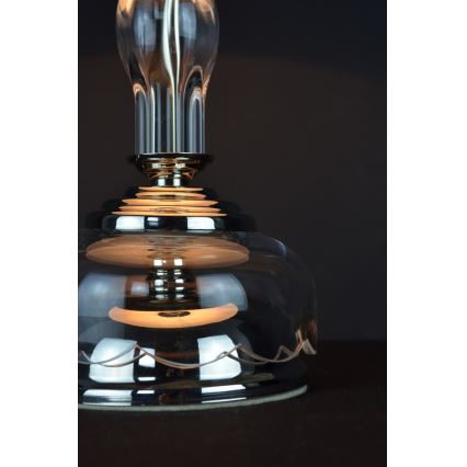 Sklenená stolová lampa s kovovou základňou svieti. Je umiestnená na tmavom povrchu.