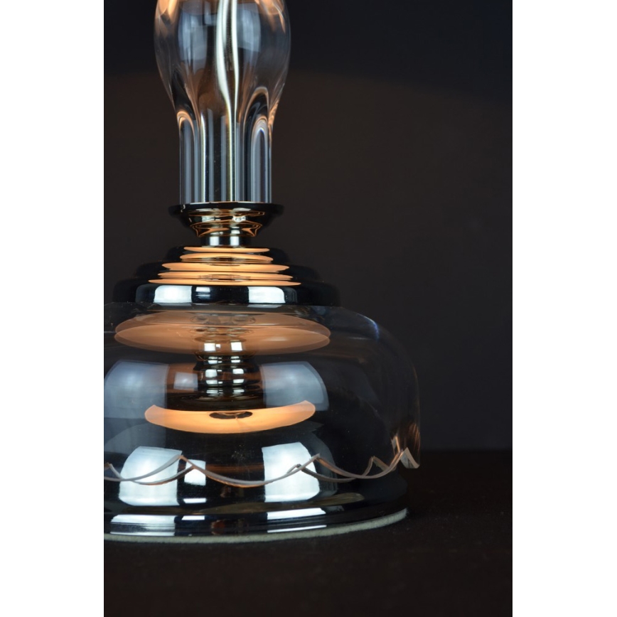 Sklenená stolová lampa s kovovou základňou svieti. Je umiestnená na tmavom povrchu.