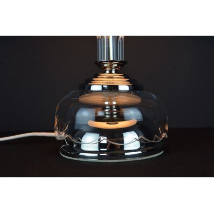 Stolová lampa svieti. Je vyrobená z priehľadného skla a chrómovaného kovu, má zvlnený okraj tienidla. Je umiestnená na tmavom povrchu.