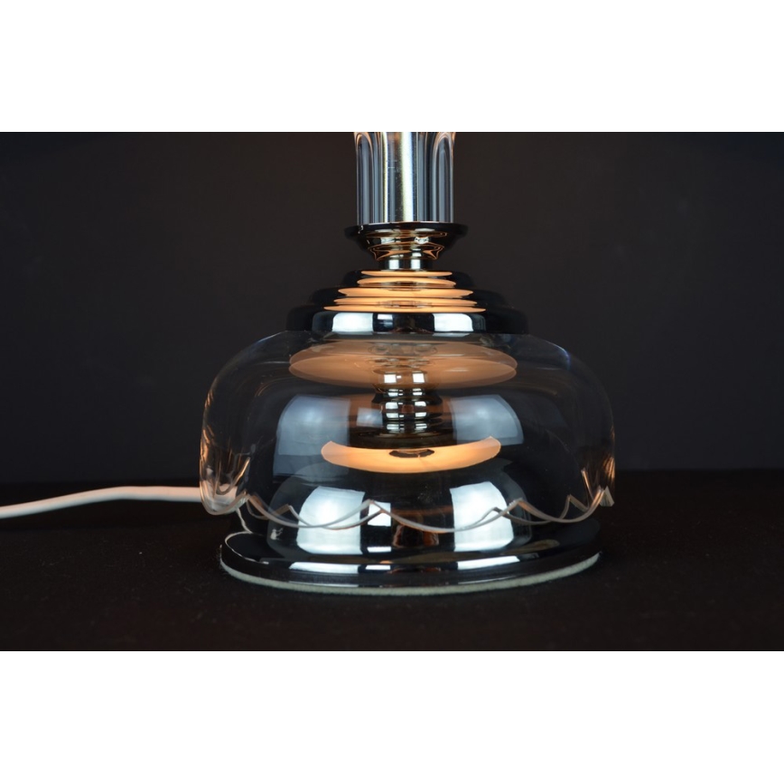 Stolová lampa svieti. Je vyrobená z priehľadného skla a chrómovaného kovu, má zvlnený okraj tienidla. Je umiestnená na tmavom povrchu.