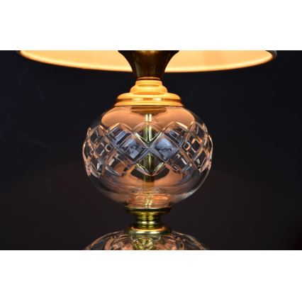 Stolová lampa s tienidlom svieti. Jej stredná časť je guľová, z číreho brúseného skla s geometrickým vzorom, spojená mosadznými prvkami. Spodná časť je tiež guľová, z brúseného skla. Tmavé pozadie.