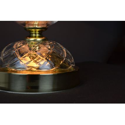 Stolová lampa svieti. Je vyrobená z priehľadného, fasetovaného skla a mosadze. Svieti na tmavom podklade.