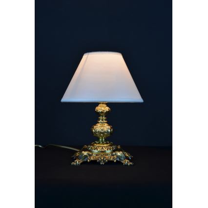 Stolová lampa svieti. Má zlatú, zdobenú základňu a biely tienidlo. Stojí na tmavom povrchu.