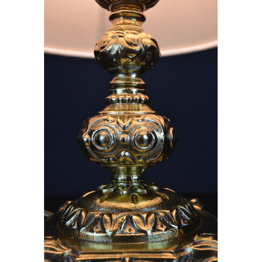 Dekoratívna stolová lampa so zlatou kovovou základňou s bohatou ornamentálnou výzdobou nesie tienidlo. Základňa je zdobená reliéfnymi motívmi listov a kvetov. Lampa stojí na tmavom podklade.