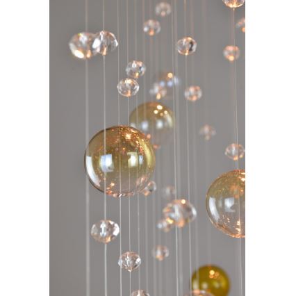 Sklenená lampa visí zo stropu. Z tenkých vlákien sú zavesené rôzne veľké sklené gule, niektoré priehľadné, iné žltohnedé, a menšie sklené kryštáliky. Svetlo presvetľuje gule.