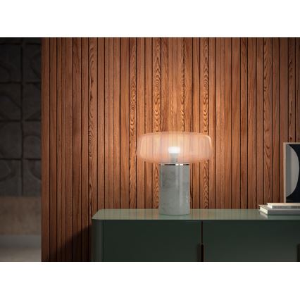 Stolová lampa s mramorovým podstavcom a priesvitným tienidlom svieti na tmavozelenej komode pred drevenou stenou.