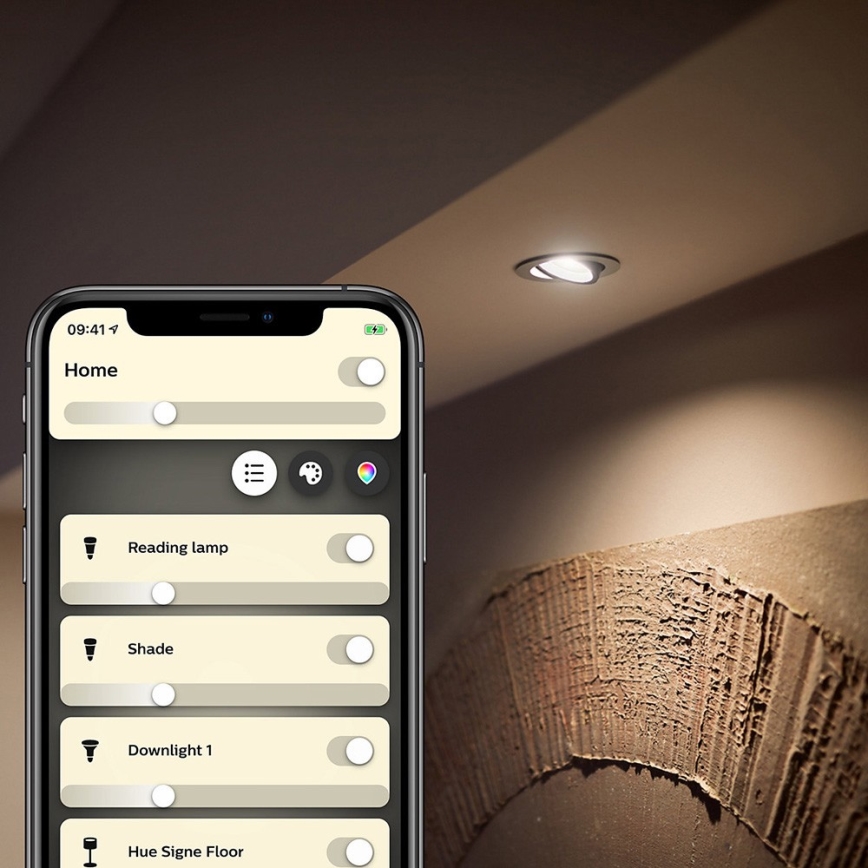 Mobilný telefón zobrazuje aplikáciu na ovládanie osvetlenia. Aplikácia umožňuje zapínať a regulovať intenzitu osvetlenia: Reading lamp, Shade, Downlight 1 a Hue Signe Floor. Na strope svieti zabudované bodové svetlo.