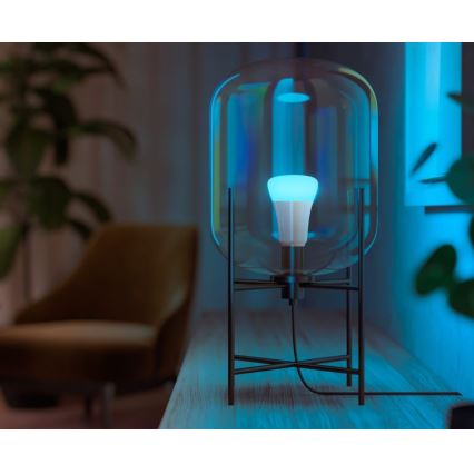Stolová lampa s objemným skleneným tienidlom svieti modrým svetlom. Lampa stojí na drevenom povrchu v tmavej miestnosti pri kresle.