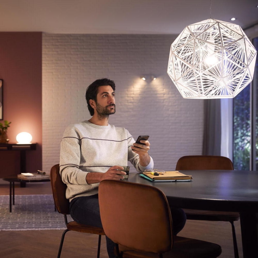 Muž sedí pri stole, drží mobilný telefón a pohár. Na stole sú knihy a okuliare. Nad stolom visí veľká geometrická lampa.