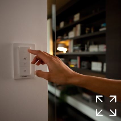 Ruka ovláda svetelný vypínač Philips na stene. Vypínač má tlačidlá pre zapínanie/vypínanie, nastavenie jasu a farebnej teploty. Pozadie tvorí rozostrená knižnica.