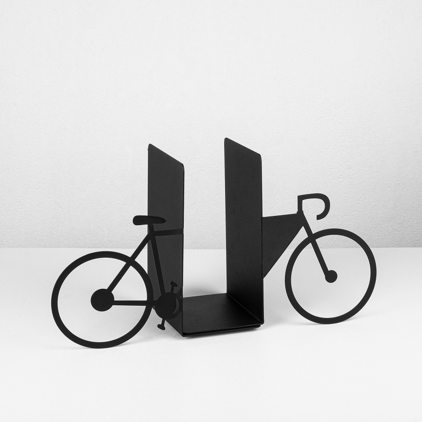 Držiak na knihy v tvare bicykla drží knihy. Je vyrobený z čiernej kovu. Stojí na bielom povrchu pred bielou stenou.