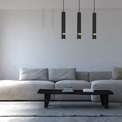 Tri čierne závesné lampy s zlatými prstencami visia nad svetlosivou pohovkou v modernom interiéri. Na nízkom drevenom konferenčnom stolíku ležia knihy.