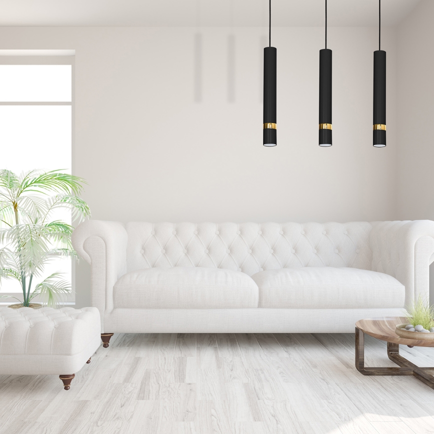 Biela pohovka stojí v svetlej miestnosti s drevenou podlahou. Nad pohovkou visia tri čierne závesné svietidlá so zlatými detailmi. Vedľa pohovky je biely taburet.