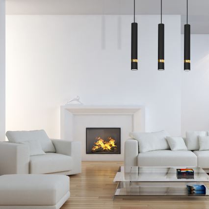 Tri čierne závesné lampy s zlatými detailmi visia nad bielou sedačkou a kreslom v obývacej izbe s bielym krbom a horiacim ohňom. Na podlahe je sklenený konferenčný stolík.